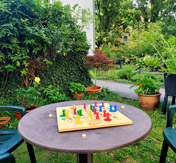 Mensch ärgere dich nicht - Spiel auf einem Gartentisch in grünem Garten mit Häusern im Hintergrund